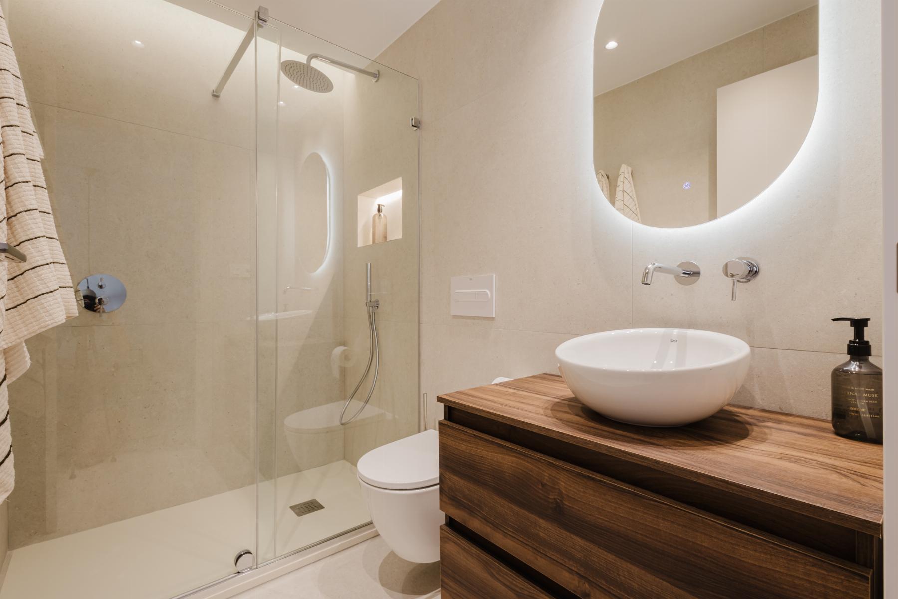Modern beige tiled bathroom with glass shower enclosure, round white vessel sink on a wooden vanity, and a backlit curved mirror.