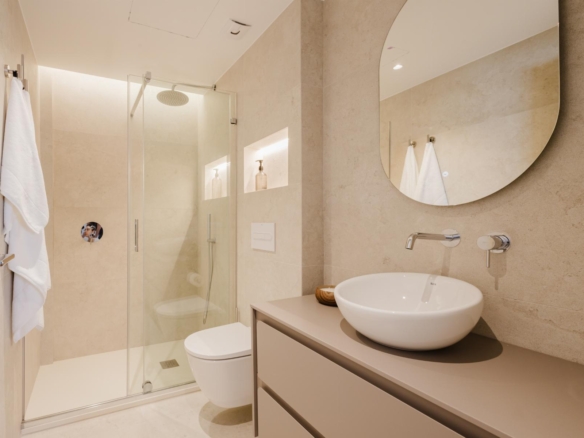 Modern bathroom with glass shower enclosure, wall-mounted faucet, and a round vessel sink under an oval mirror; beige tiled walls and white towels.