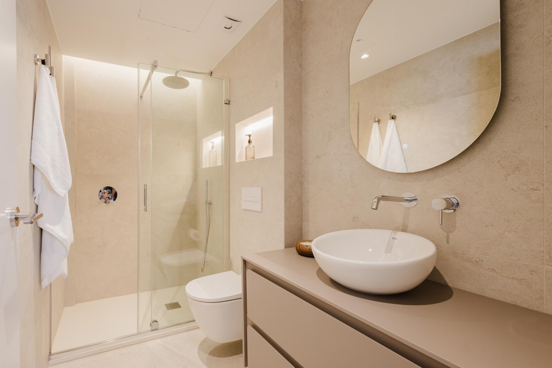 Modern bathroom with glass shower enclosure, wall-mounted faucet, and a round vessel sink under an oval mirror; beige tiled walls and white towels.