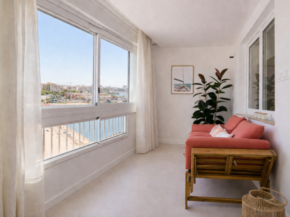 Bright coastal living area with a coral sofa and large window overlooking the waterfront town and sea beyond, white walls and sheer curtains, plant in corner.