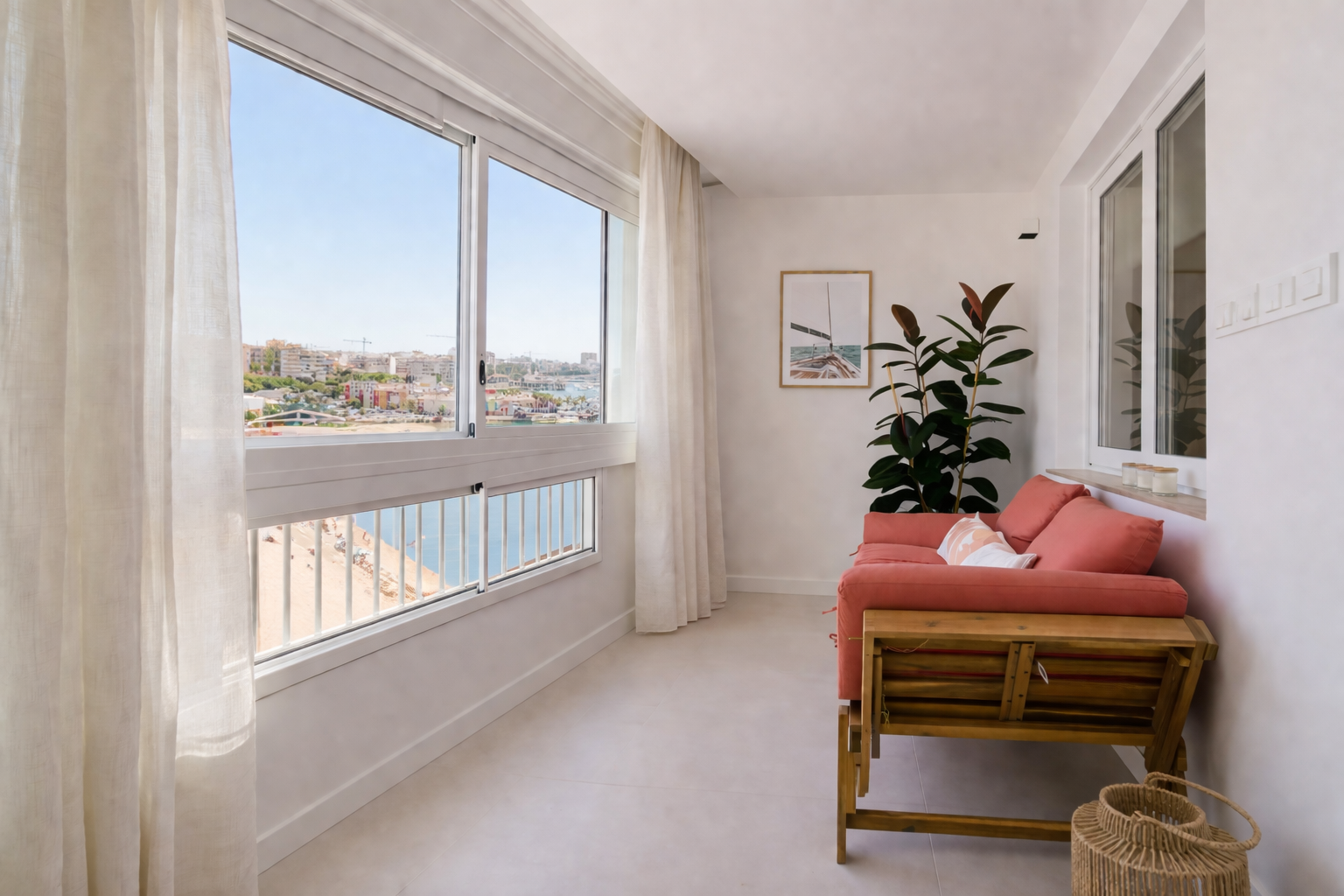 Bright coastal living area with a coral sofa and large window overlooking the waterfront town and sea beyond, white walls and sheer curtains, plant in corner.