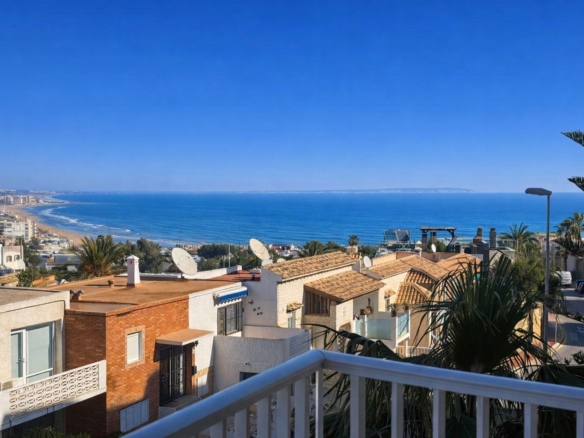 Coastal hillside neighborhood with white and tan houses overlooking a blue ocean and sandy beach under a clear sky.