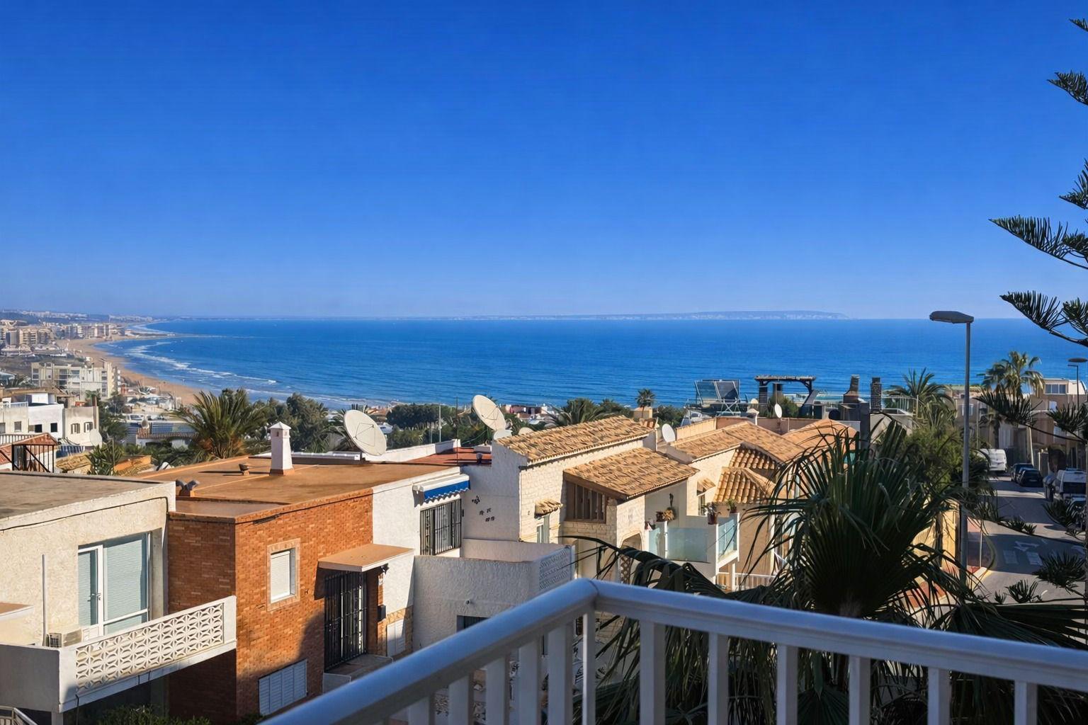 Coastal hillside neighborhood with white and tan houses overlooking a blue ocean and sandy beach under a clear sky.