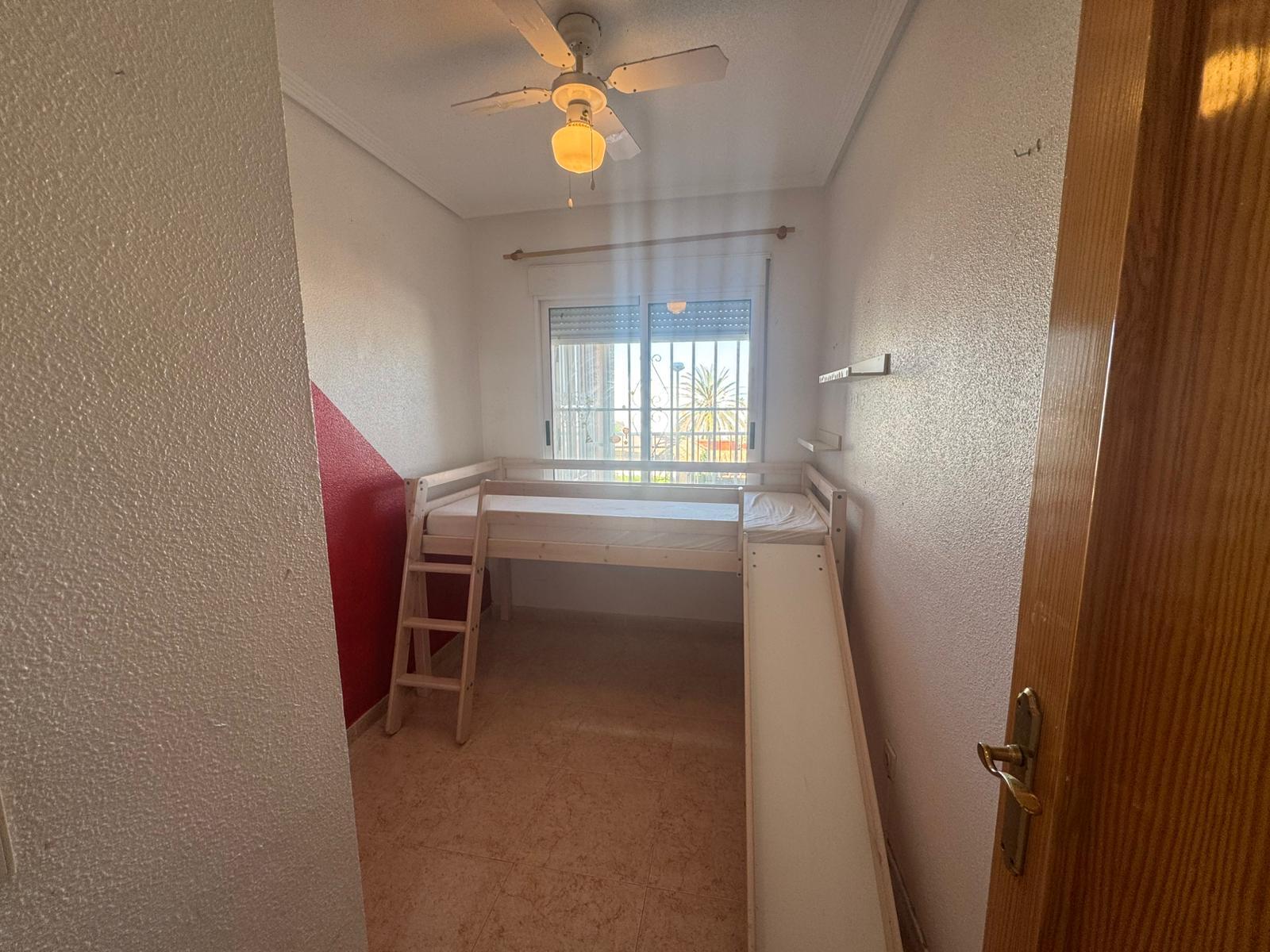 Narrow bedroom with a white bunk bed, a built-in slide on the right, and a wooden ladder on the left; red diagonal accent on the left wall, a window with bars at the far end, and a ceiling fan above.