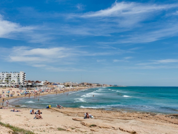 Busy sandy beach with sunbathers and families, blue sea to the right and low-rise hotels along the shore under a clear sky.