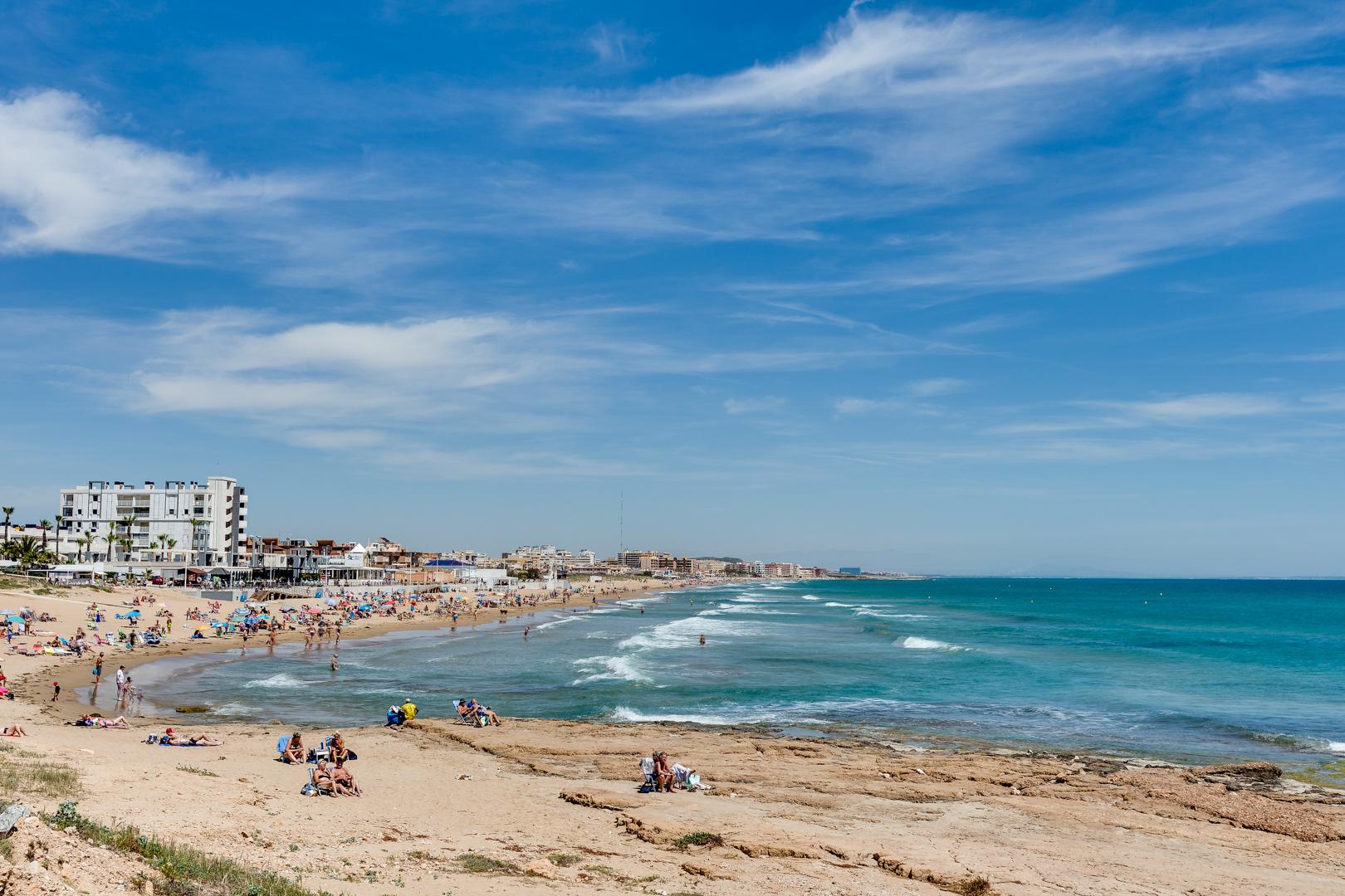 Busy sandy beach with sunbathers and families, blue sea to the right and low-rise hotels along the shore under a clear sky.