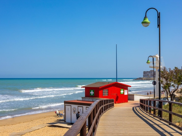 Sunny beach boardwalk with a red lifeguard hut beside the sea and green streetlamps under a clear blue sky.