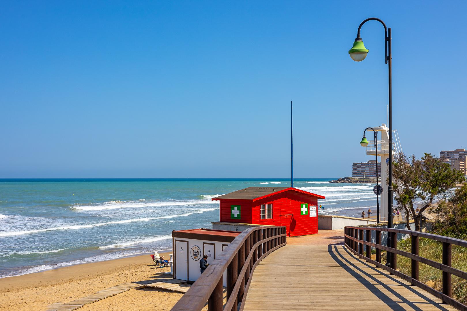Sunny beach boardwalk with a red lifeguard hut beside the sea and green streetlamps under a clear blue sky.