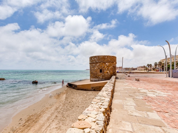 Circular stone tower at a seawall along a sandy beach, with calm blue sea and a paved promenade.
