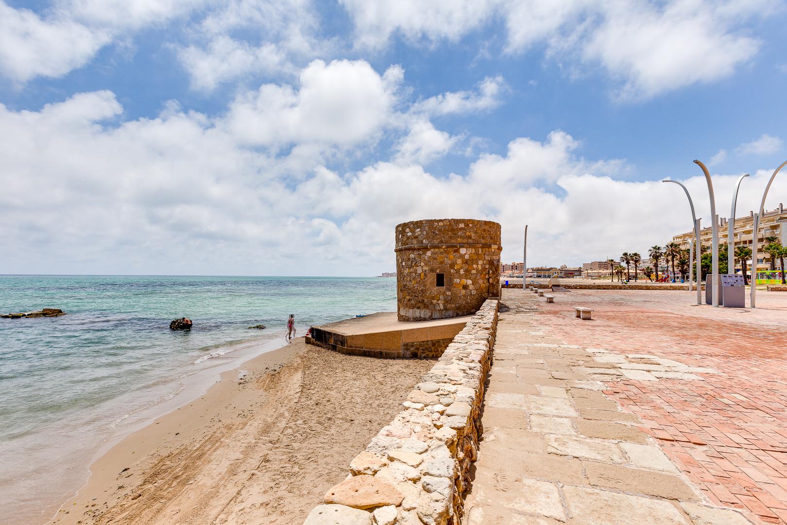 Circular stone tower at a seawall along a sandy beach, with calm blue sea and a paved promenade.