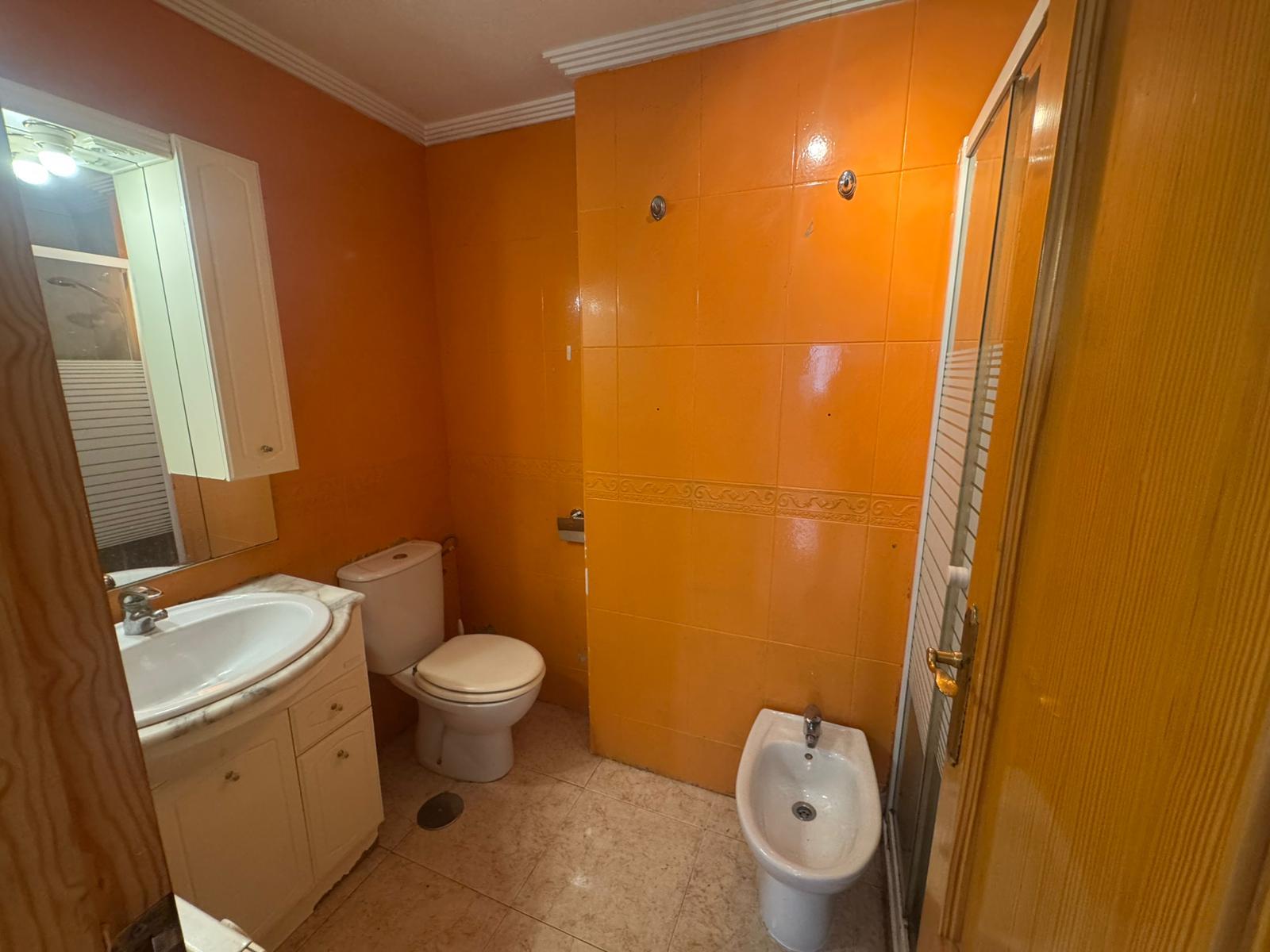 Small bathroom with orange tiled walls, white sink and vanity, toilet, and a bidet near the door; shower enclosure on the right.