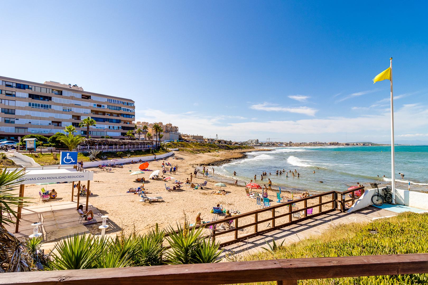 Busy sandy beach along a coastal town with apartment buildings, palm trees, and people sunbathing under a bright sky.