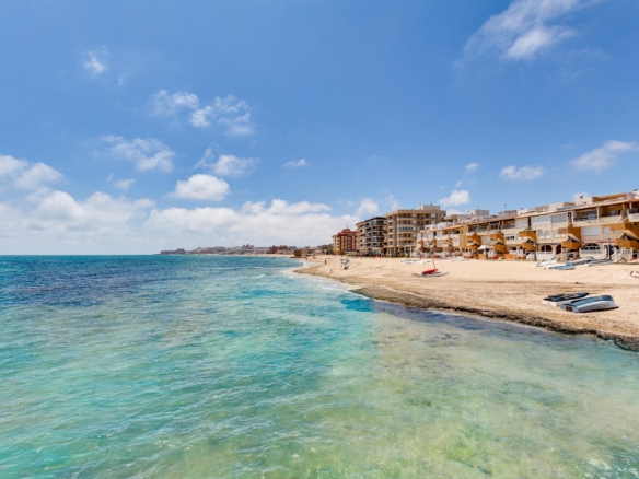 Sunny coastal scene with a sandy beach, turquoise sea, and a row of seaside apartments along the shore under a blue sky.