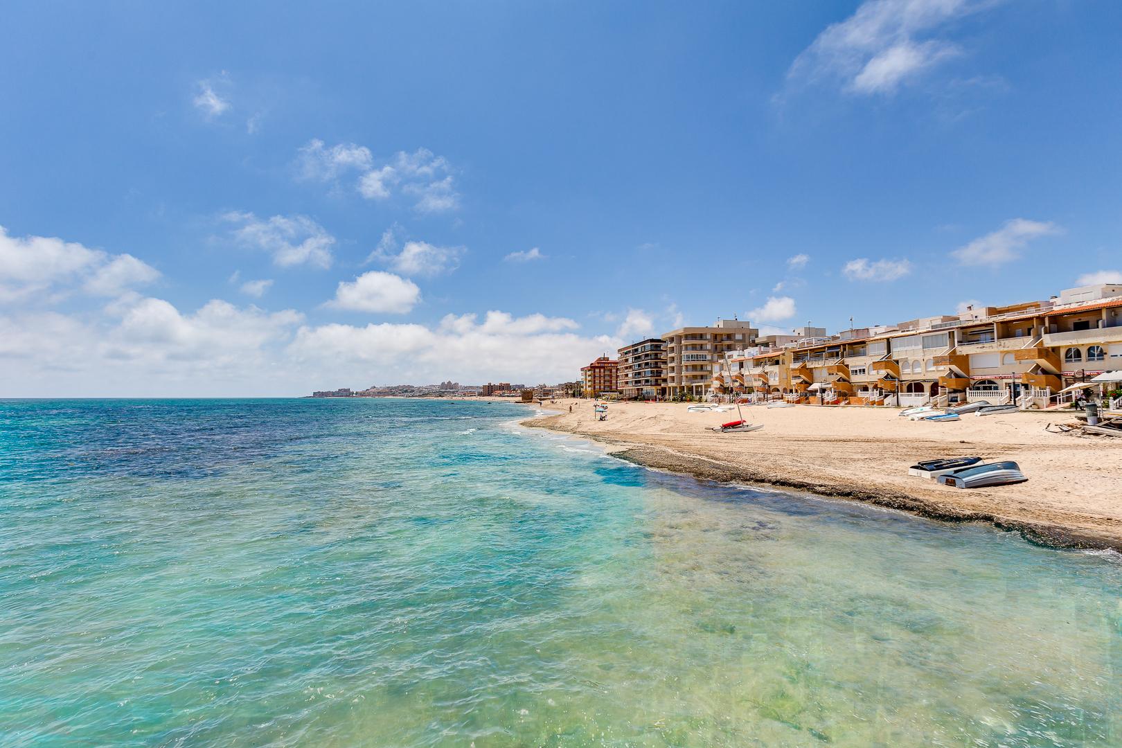 Sunny coastal scene with a sandy beach, turquoise sea, and a row of seaside apartments along the shore under a blue sky.