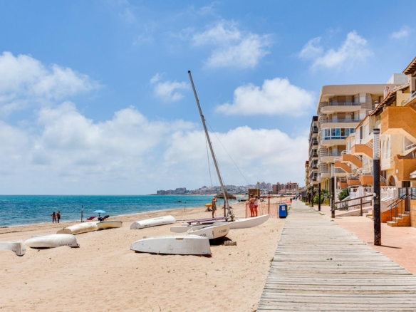 Beach scene with several small boats on the sand, a wooden boardwalk, and modern orange apartment buildings along the shore under a blue sky.
