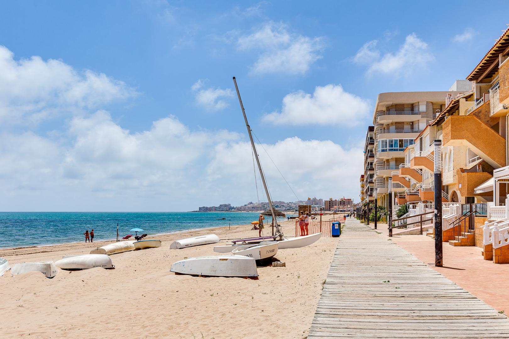 Beach scene with several small boats on the sand, a wooden boardwalk, and modern orange apartment buildings along the shore under a blue sky.