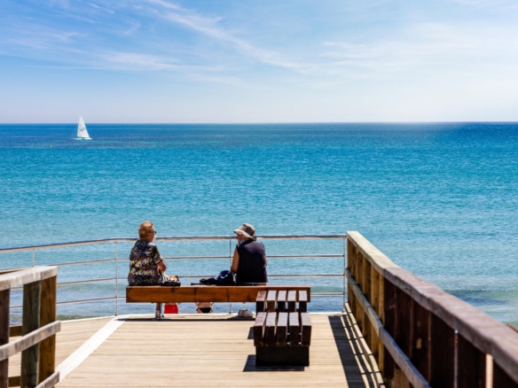 Two people sit on a wooden bench on a seaside pier, looking out over the calm blue ocean with a small sailboat on the horizon.