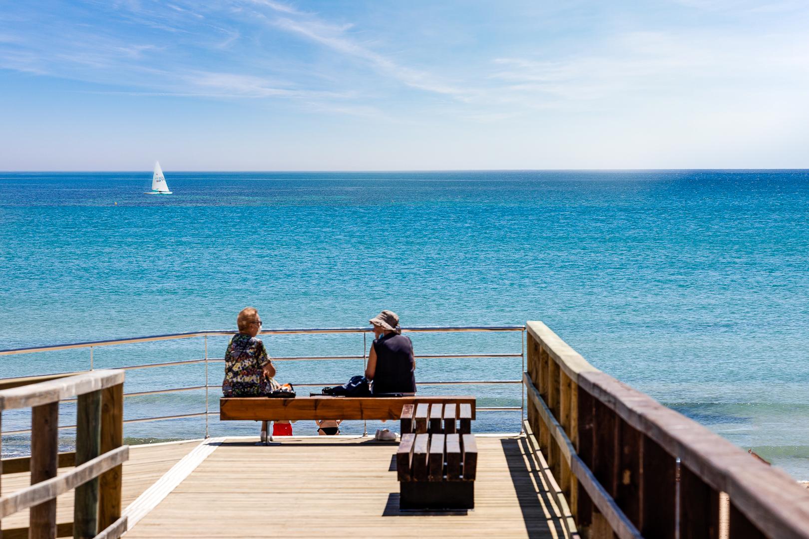 Two people sit on a wooden bench on a seaside pier, looking out over the calm blue ocean with a small sailboat on the horizon.