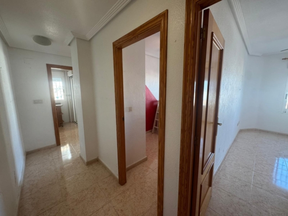 Interior hallway with multiple open doors, wooden door frames, white walls, and tiled floor; glimpses of a red-walled stair area in the background