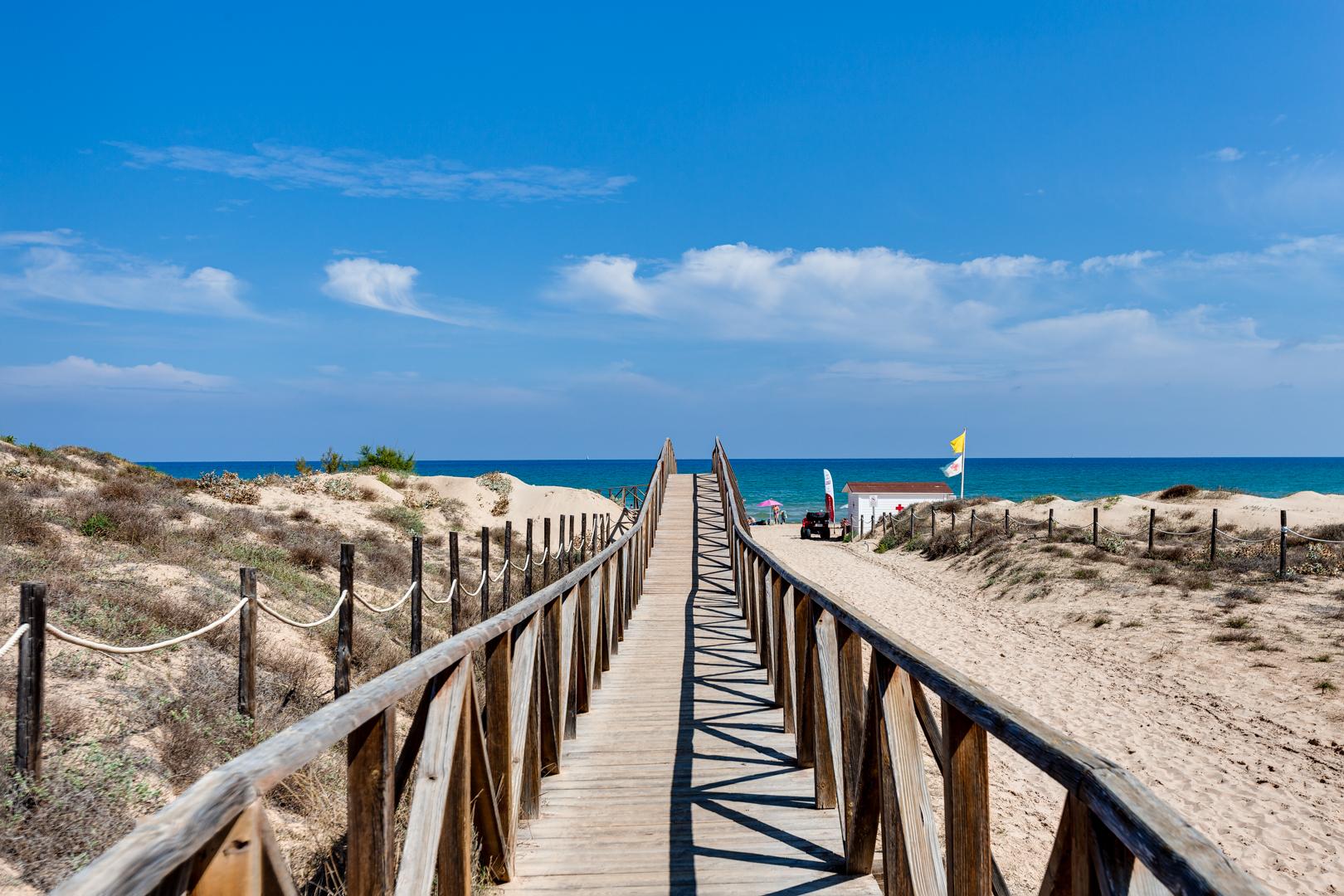 Wooden boardwalk stretching over sandy dunes toward a blue ocean under a clear sky as far as the eye can see.