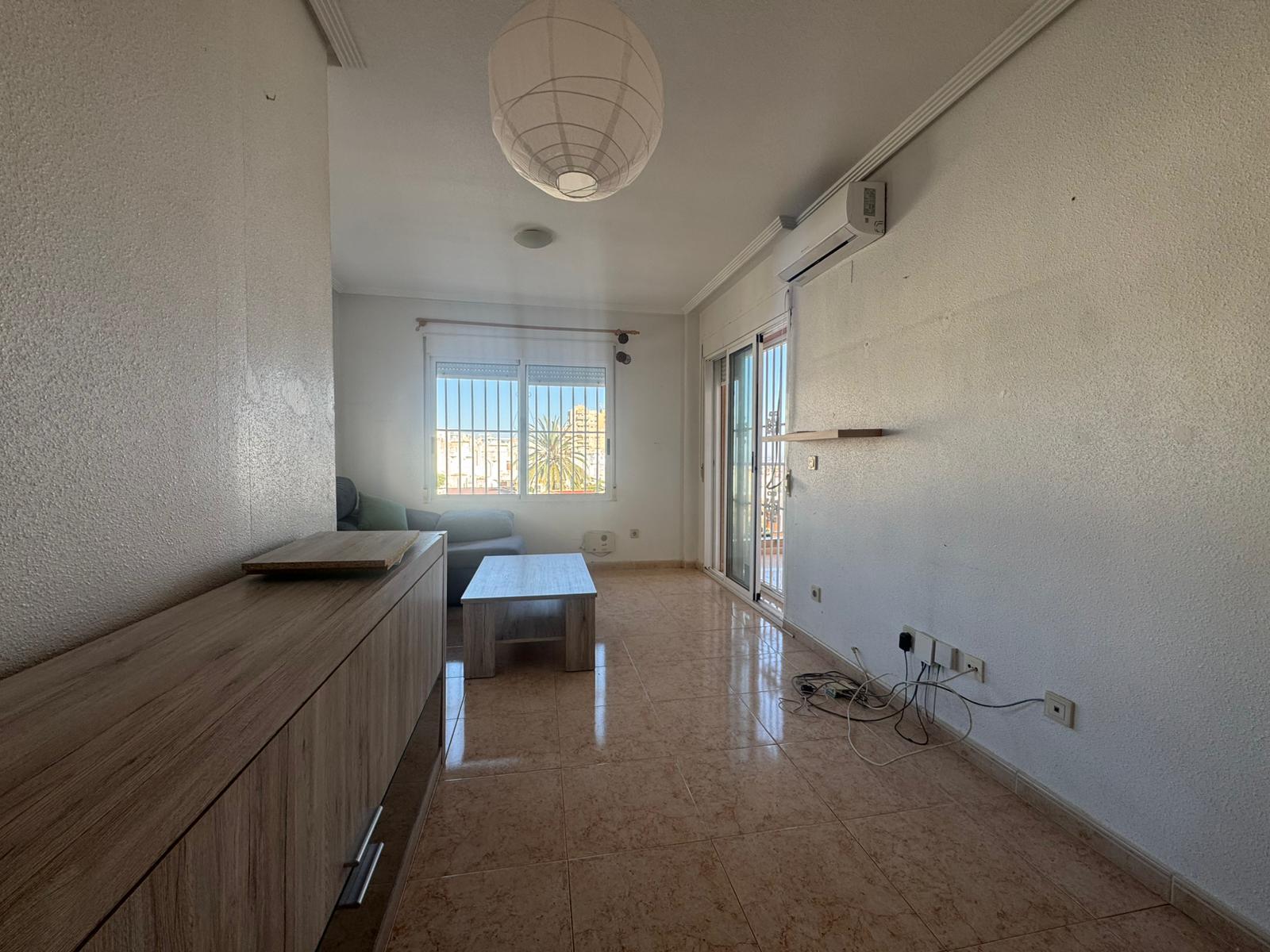 Living room with a wooden cabinet along the left wall, a light coffee table, and a gray sofa by a window with bars; balcony door on the right; tangled cables on the floor near outlets.