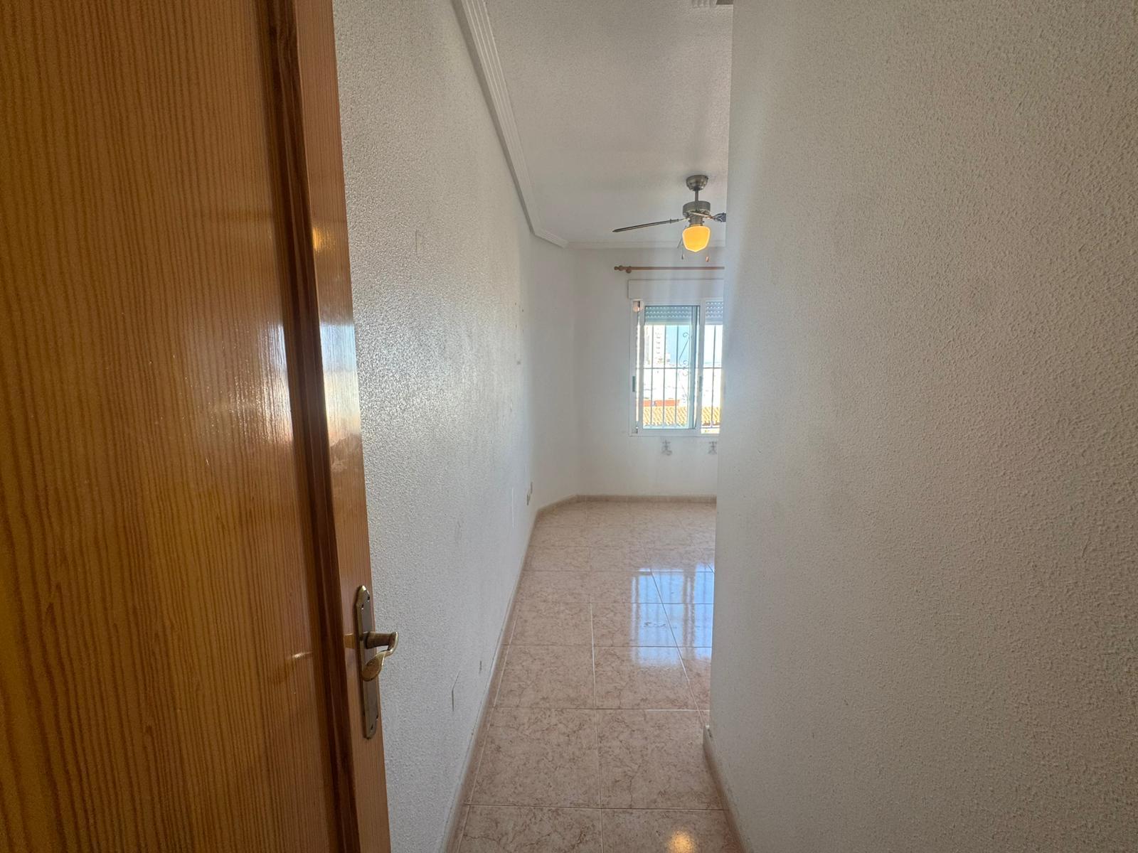 Narrow hallway with textured white walls, a wooden door on the left, and a ceiling fan; window at the far end with bars.