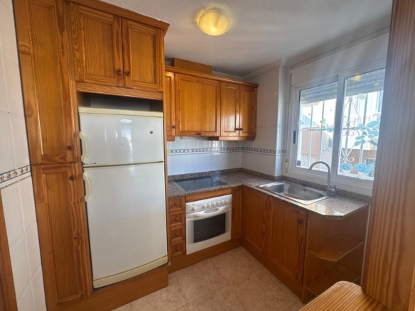 Oak‑cabinet kitchen with a white fridge, stove, and granite countertops under a ceiling light, adjacent to a window over the sink.