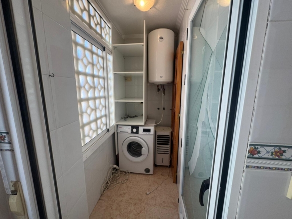 Narrow utility room with a front-loading washing machine, tall white water heater, and open shelves above. Cords lie on the tiled floor.