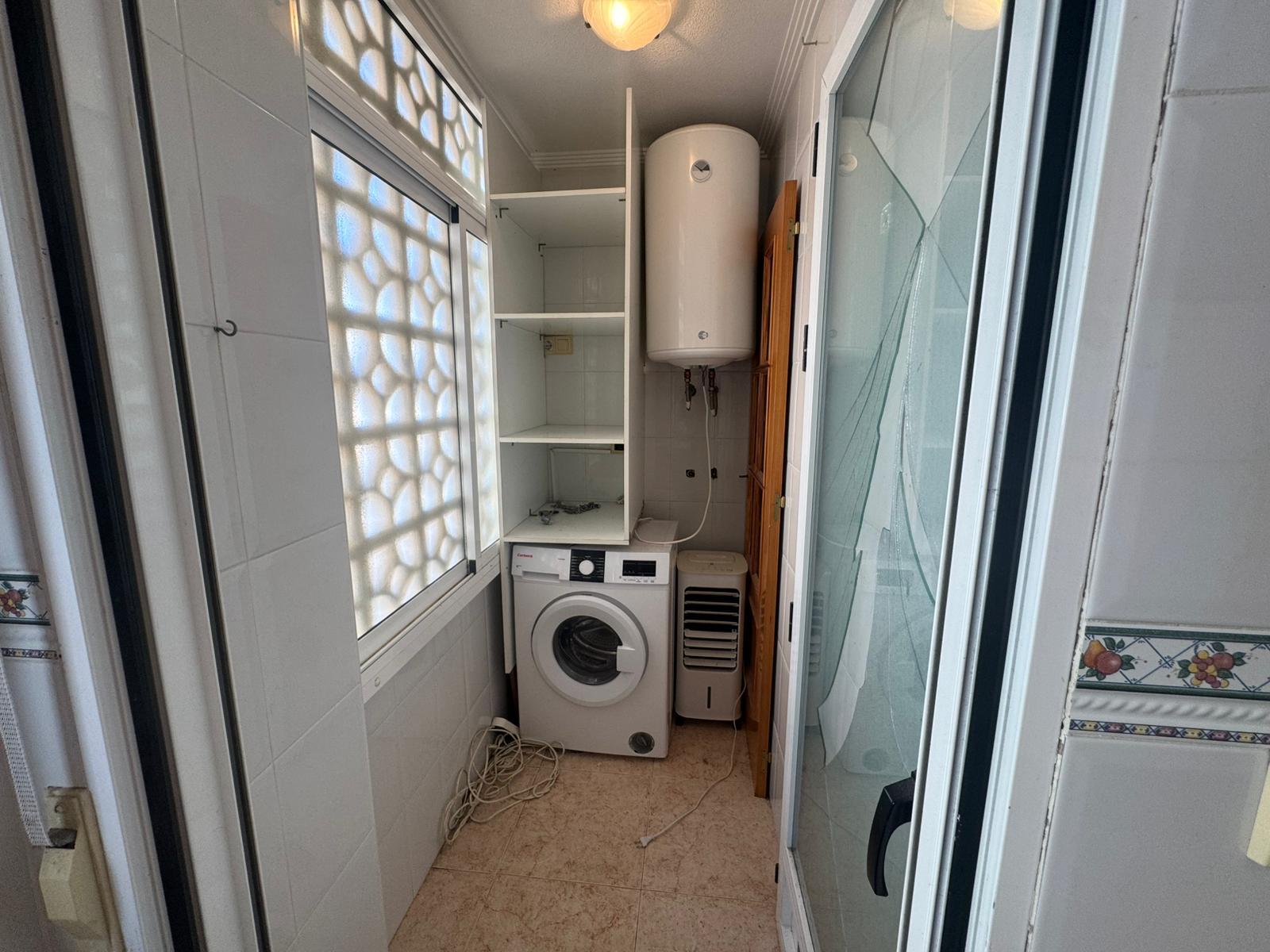 Narrow utility room with a front-loading washing machine, tall white water heater, and open shelves above. Cords lie on the tiled floor.