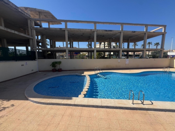 Outdoor pool area with a large unfinished concrete building frame nearby, under a clear blue sky with palm trees in the background.