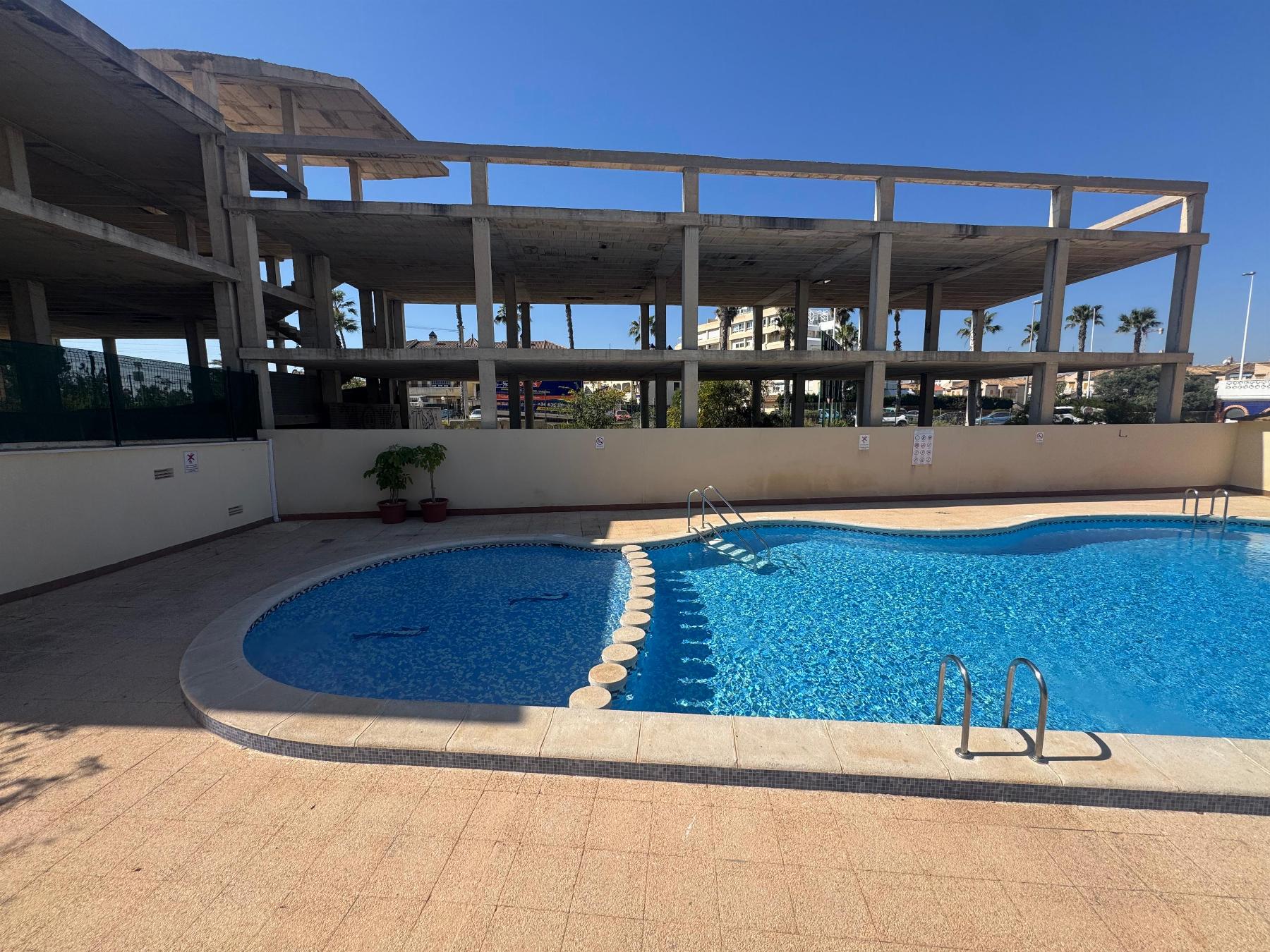 Outdoor pool area with a large unfinished concrete building frame nearby, under a clear blue sky with palm trees in the background.