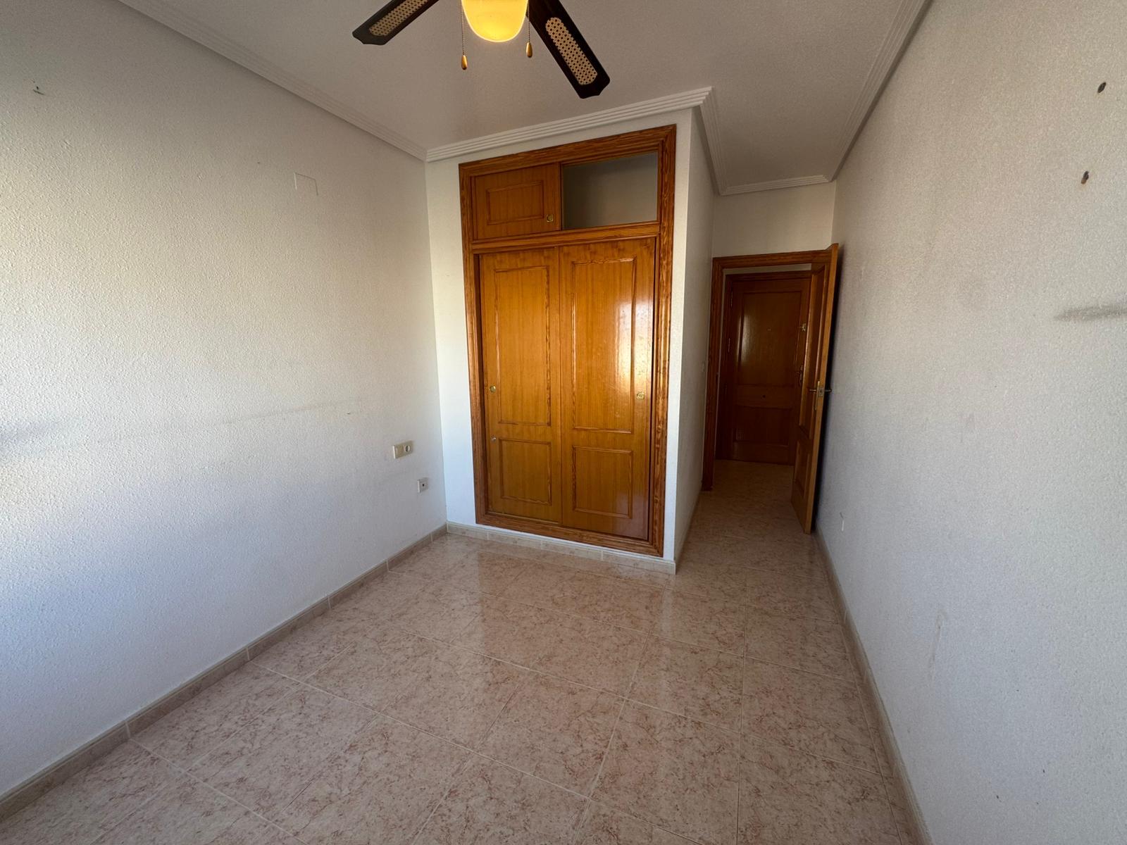 Empty small room with beige tiled floor, white walls, a wooden built-in wardrobe, and a ceiling fan; door to a hallway on the right.