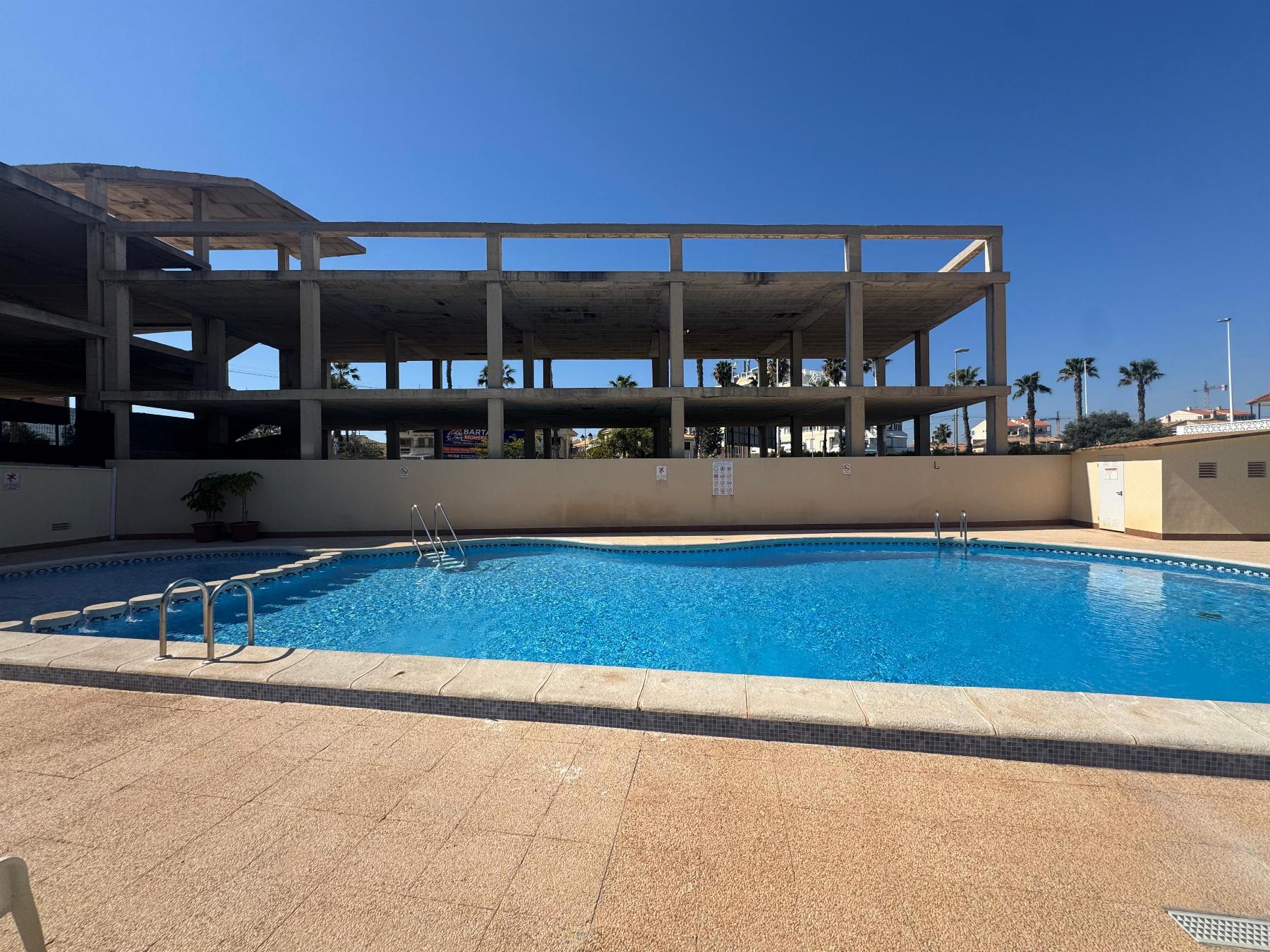 Empty outdoor swimming pool with metal ladders in the foreground and an unfinished concrete building behind under a clear blue sky.