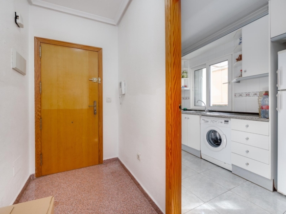 Hallway with a wooden door on the left opening into a bright white kitchen featuring a washing machine beneath a window.