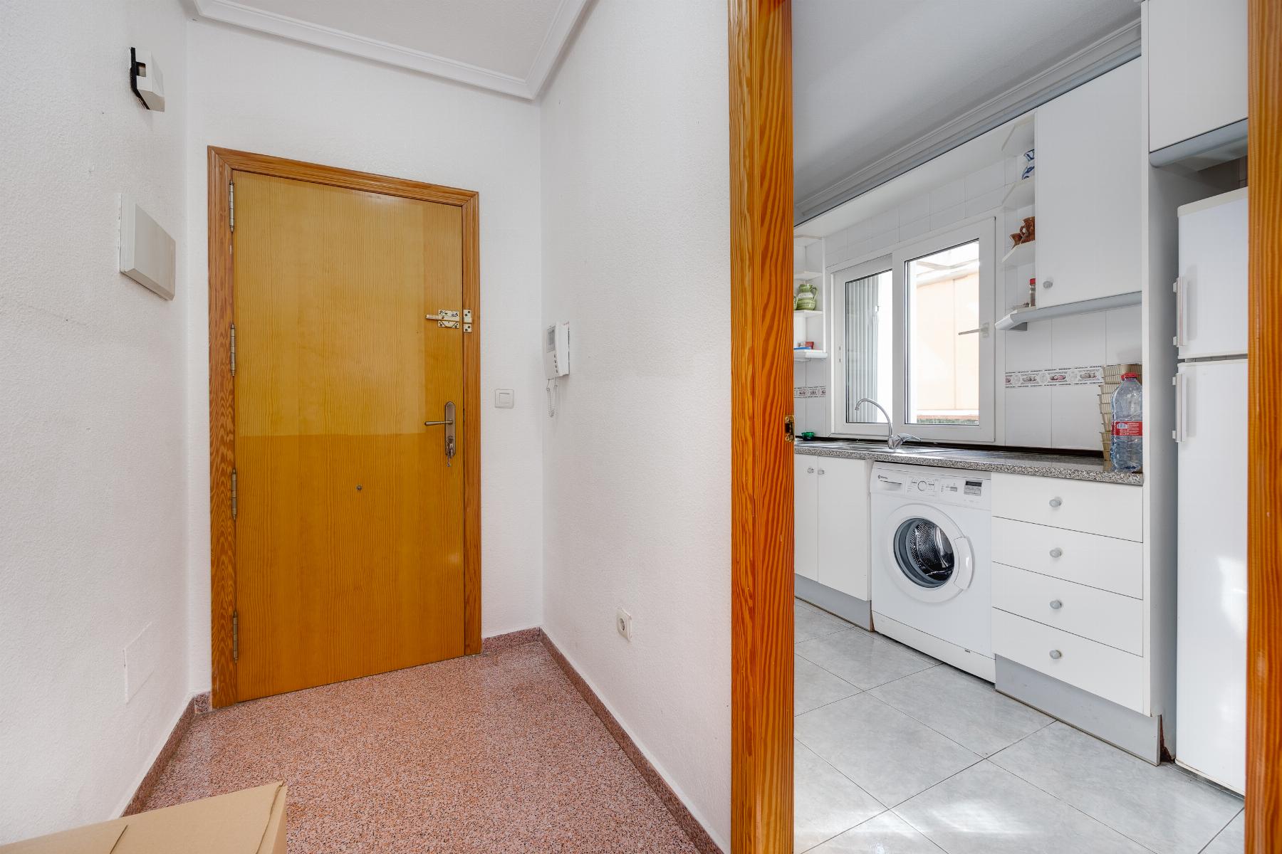 Hallway with a wooden door on the left opening into a bright white kitchen featuring a washing machine beneath a window.