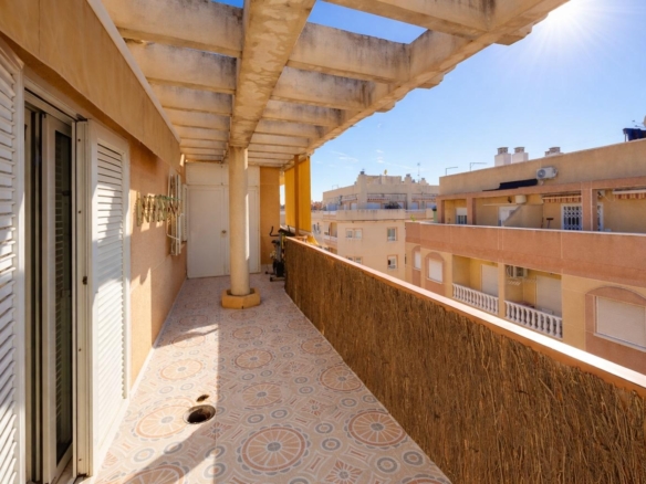 Sunlit apartment balcony with patterned tile floor, white shutters, and a long railing facing city buildings under a wooden pergola.