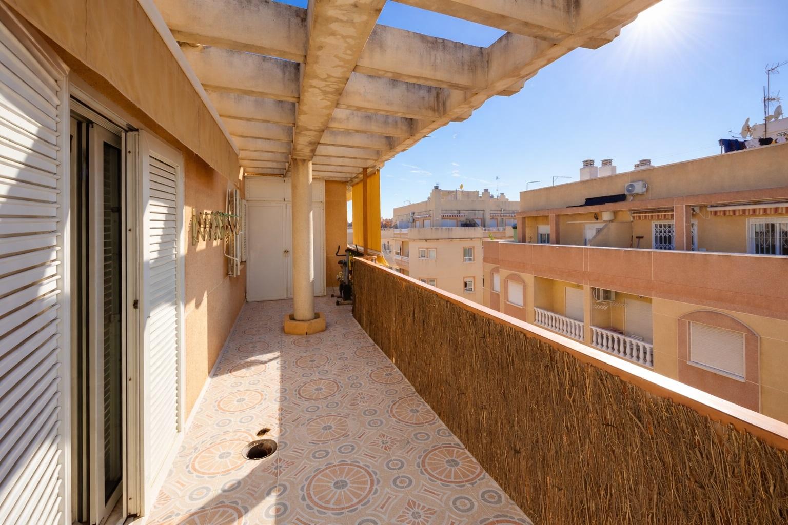 Sunlit apartment balcony with patterned tile floor, white shutters, and a long railing facing city buildings under a wooden pergola.