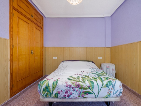 Small bedroom with lavender walls, wood-paneled lower walls, a single bed with a floral duvet, a round ceiling light, and a large wooden wardrobe on the left.