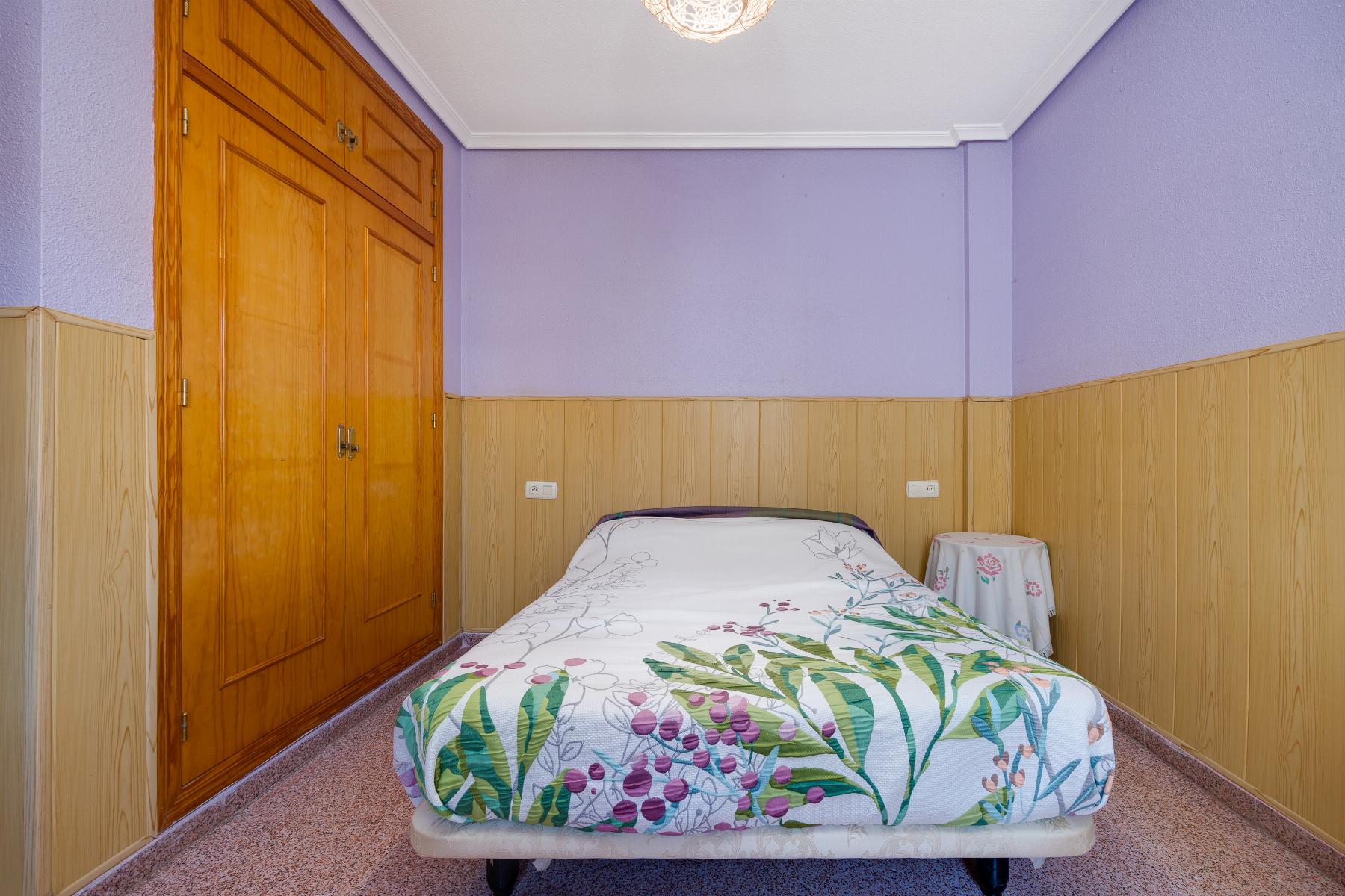 Small bedroom with lavender walls, wood-paneled lower walls, a single bed with a floral duvet, a round ceiling light, and a large wooden wardrobe on the left.