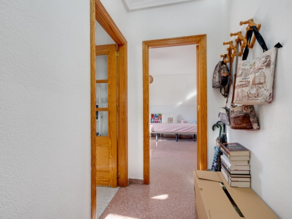 Hallway with wooden door frames leads to a bedroom with a pink bedspread; bags and coats hang on the right, stacked boxes on a bench nearby.