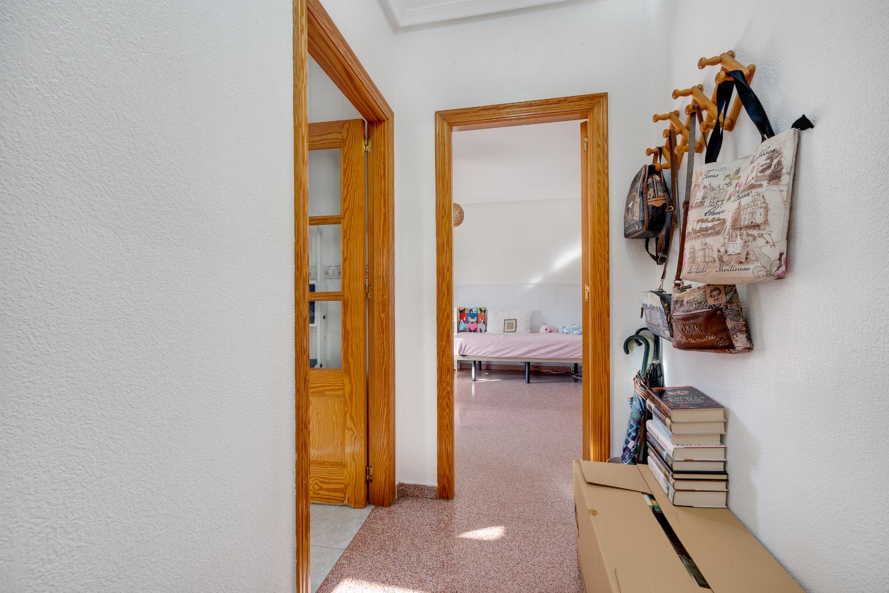Hallway with wooden door frames leads to a bedroom with a pink bedspread; bags and coats hang on the right, stacked boxes on a bench nearby.