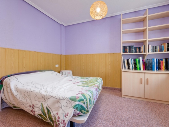 Bedroom with lavender walls, wood wainscoting, and a bed with floral quilt looking toward a tall bookcase.