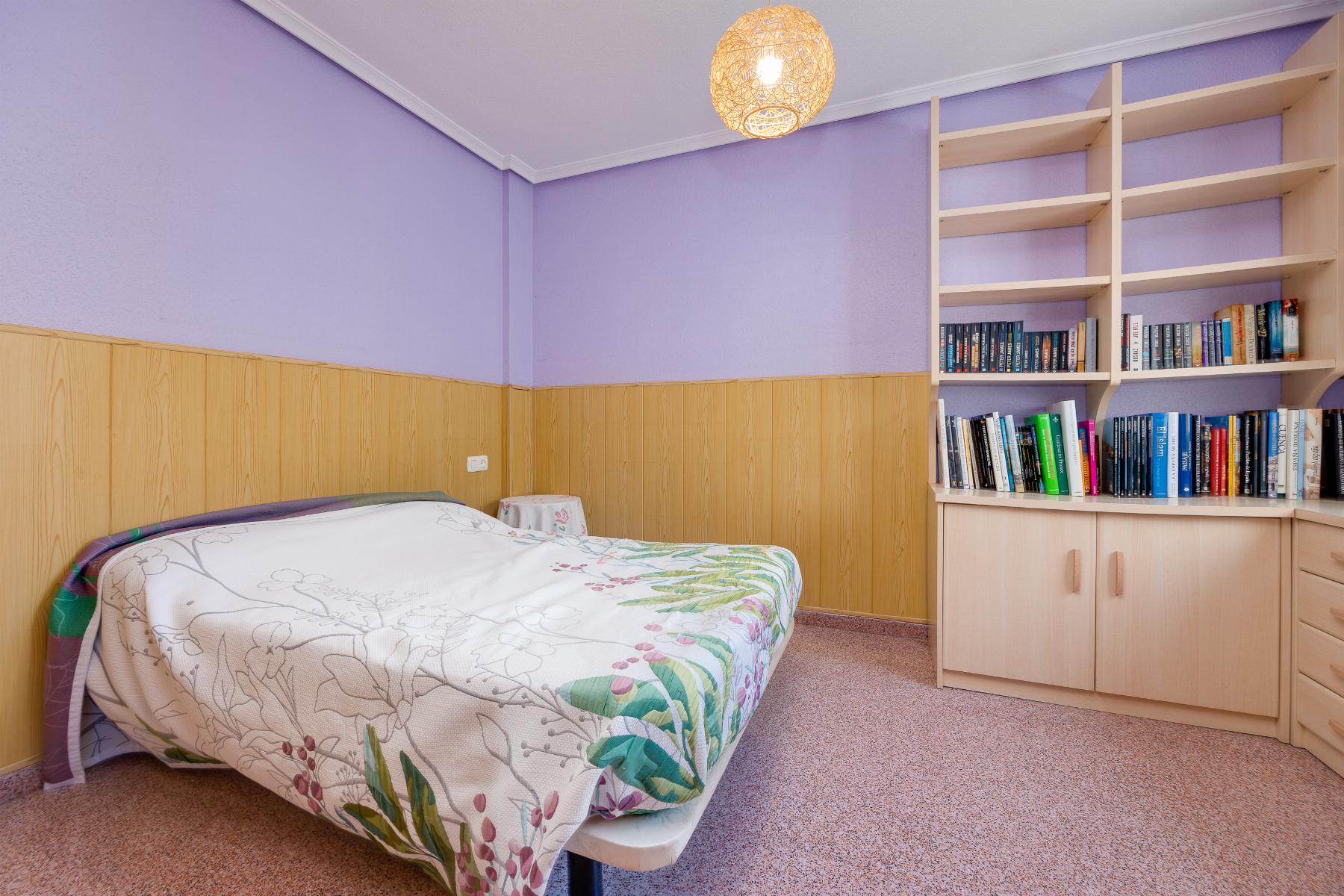 Bedroom with lavender walls, wood wainscoting, and a bed with floral quilt looking toward a tall bookcase.