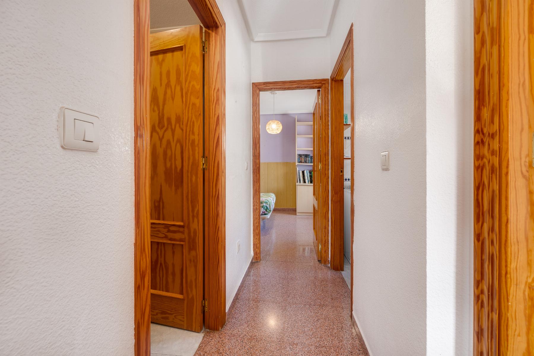 Hallway with white walls and open wooden door frames, leading to a bedroom with a bed and bookshelf.
