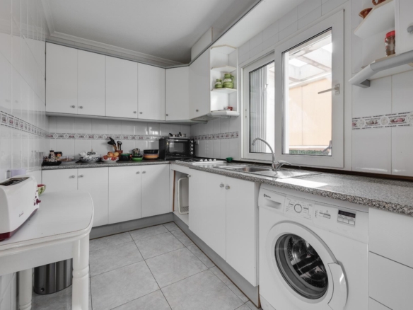 Modern white U-shaped kitchen with grey speckled countertops, upper and lower cabinets, a sink under a large window, and a front-loading washing machine nearby.