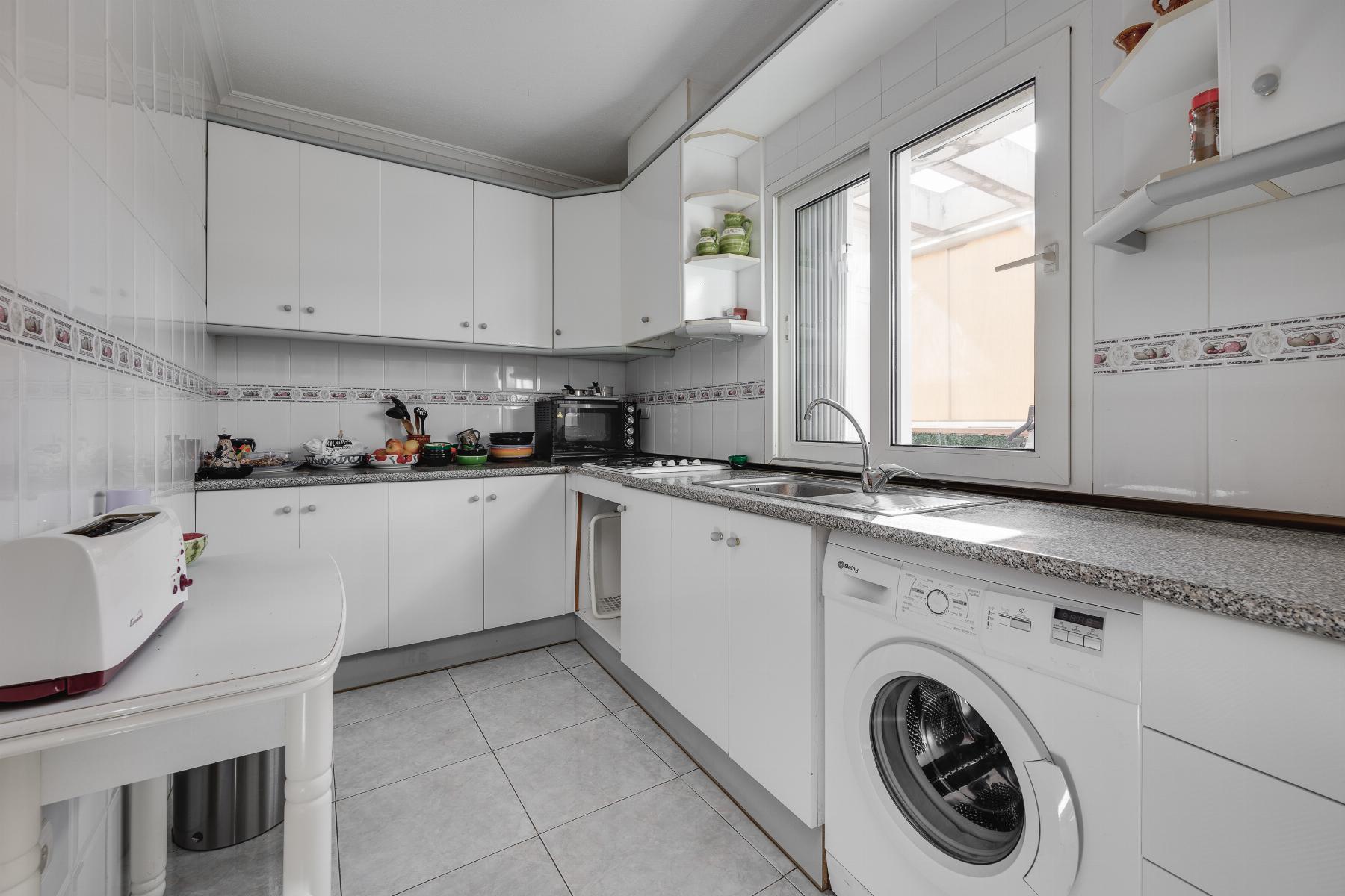 Modern white U-shaped kitchen with grey speckled countertops, upper and lower cabinets, a sink under a large window, and a front-loading washing machine nearby.