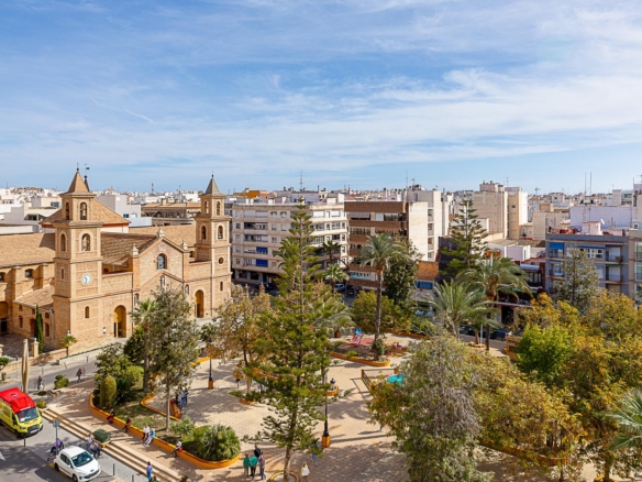 A plaza with a historic brick church, palm trees, benches, and people walking in a sunny city square.