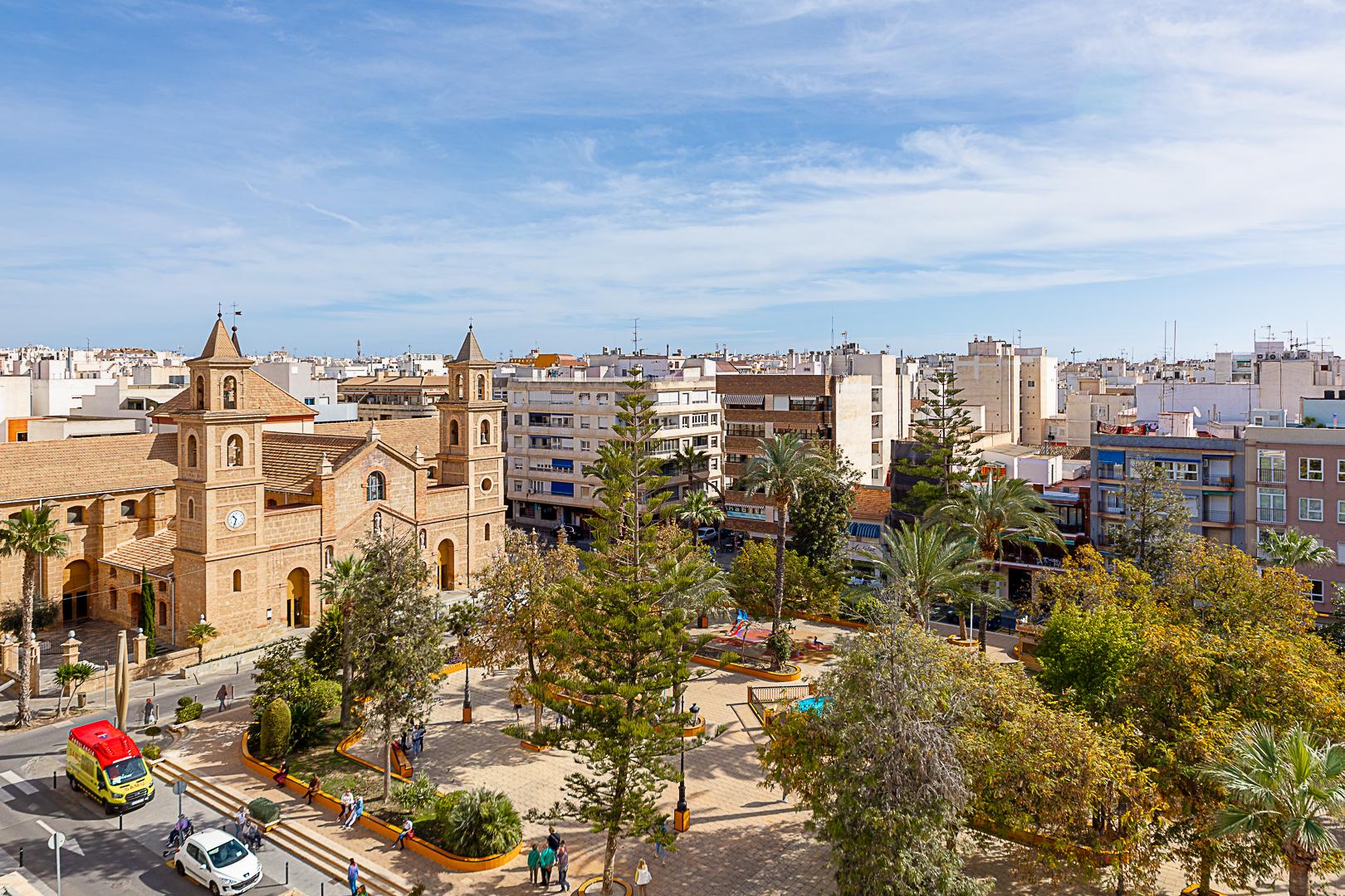 A plaza with a historic brick church, palm trees, benches, and people walking in a sunny city square.