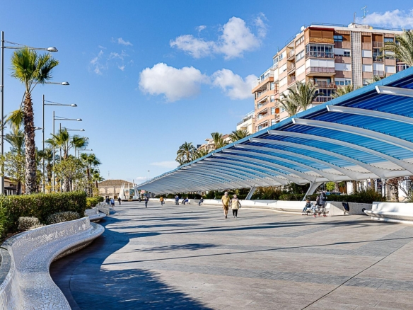 Sunlit seaside promenade with a curved blue canopy, palm trees, and people strolling along a tiled plaza next to a mosaic water channel on the left.