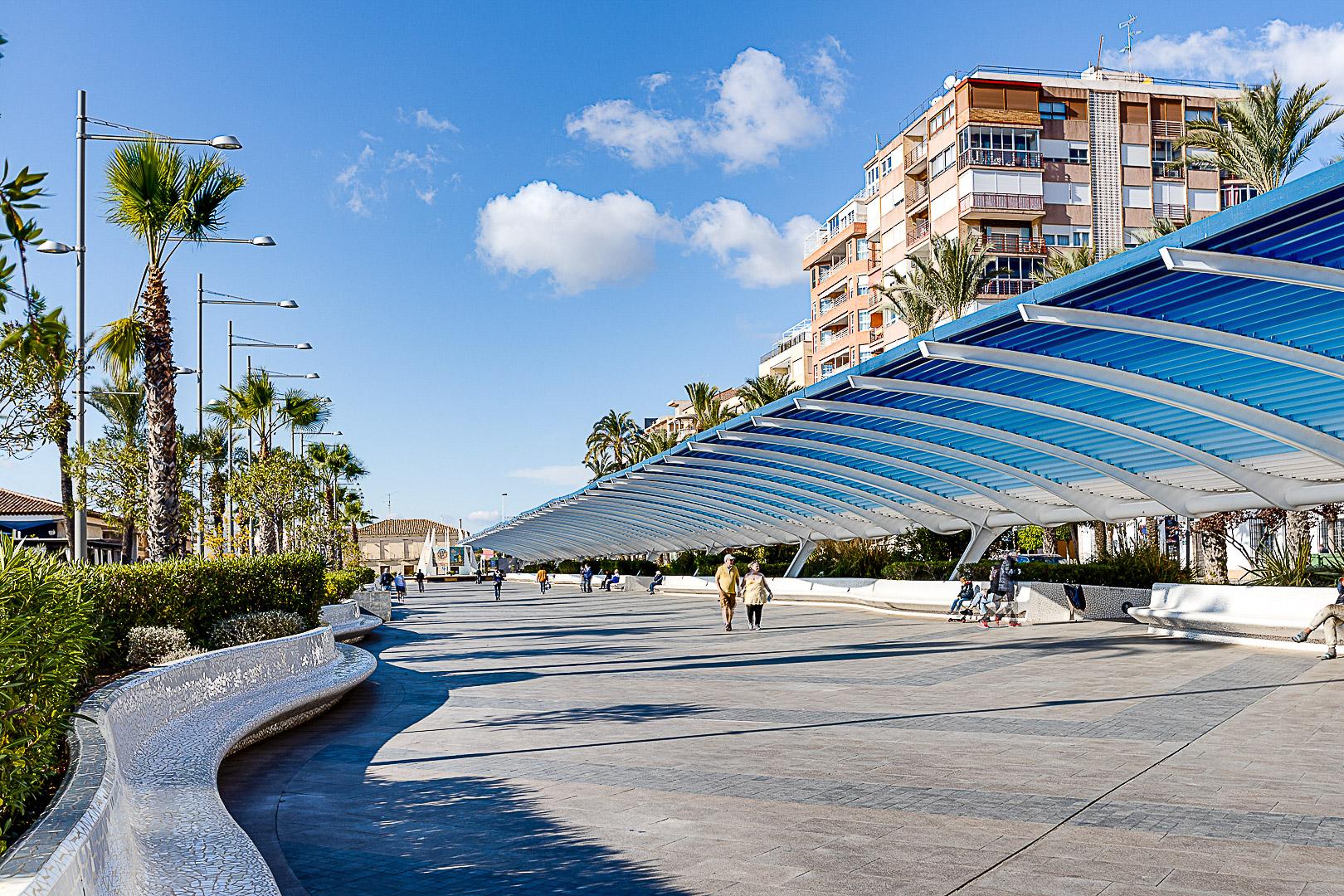 Sunlit seaside promenade with a curved blue canopy, palm trees, and people strolling along a tiled plaza next to a mosaic water channel on the left.